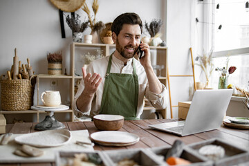 Caucasian male potter entrepreneur talking to buyer of handmade bowl sitting at table and taking order using laptop. Successful businessman owner of shop made of clay talking to customer.