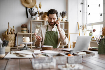 Caucasian male potter entrepreneur talking to buyer of handmade bowl sitting at table and taking order using laptop. Successful businessman owner of shop made of clay talking to customer.