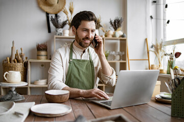 Mature Caucasian man in apron sits at table in ceramics shop and works with laptop. Experienced...