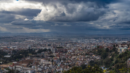 Fototapeta premium The urban landscape. Top view of the modern metropolis. Lots of densely built multi-storey buildings, green vegetation. Mountains in the distance. Picturesque storm clouds in the sky. Madagascar. 