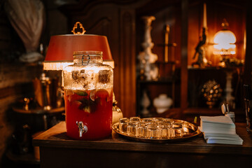 Riga, Latvia, - August 26, 2023 - A glass jar of fruit punch with lemon and lime slices, next to a tray of glasses, napkins, and a vintage lamp, creating a cozy, inviting setting.