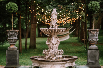 Naklejka premium Riga, Latvia, - August 26, 2023 - Ornate stone fountain with fish sculptures in a lush garden, surrounded by trees, string lights, and topiary, creating a serene and enchanting atmosphere.