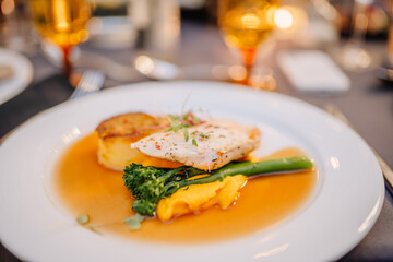 Gourmet dish with fish fillet, roasted potatoes, broccolini, and sauce, elegantly plated on a white dish with a blurred background of dining setting.