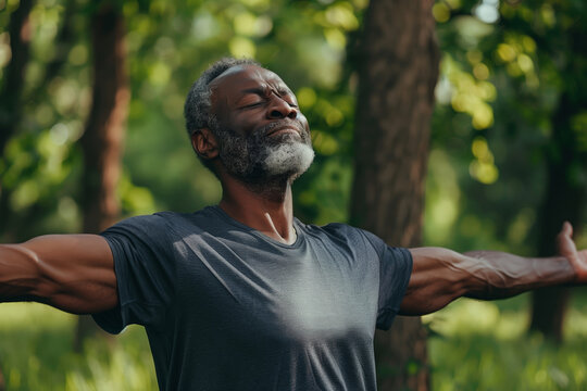 A senior man with gray hair and a beard is standing in a park, practicing breathing exercises. He has his arms outstretched and his eyes closed, enjoying the fresh air and sunshine