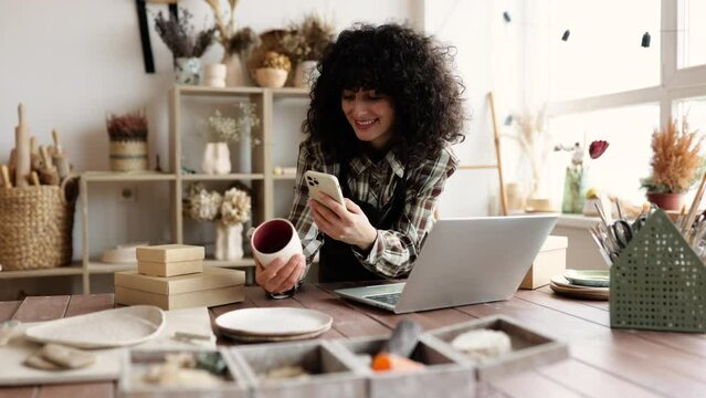 Successful girl store employee with ceramic tableware shoots photos on smartphone. Charismatic Caucasian woman seller in souvenir shop takes pictures of goods at phone for posting on social networks.