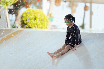 A young girl is sitting on a water slide, wearing a black shirt and goggles. The scene is playful...