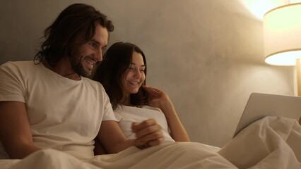 Happy couple relaxing in bed, holding hands and talking, with a laptop nearby. Cozy nighttime ambiance with warm lighting. Cinematic 4k footage, design for advertising, and social media, vlog