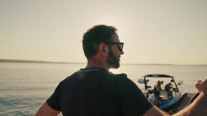 A man stands on a lake pier at sunset, leaning on a water board. Male surfer holding a short...