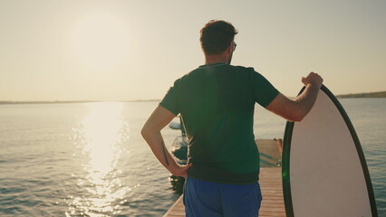 A man stands on a lake pier at sunset, leaning on a water board. Male surfer holding a short...