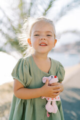 Smiling little girl hugs a toy pink mouse while standing on the seashore