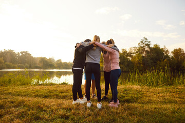Group of sporty people in sportswear standing in a circle on the lake shore hugging after workout in nature at sunset in the park. Outdoors training, team work and fitness exercises concept.