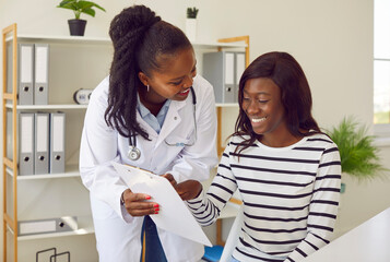 Female friendly doctor talking with a happy smiling african american woman patient sitting at the desk in office and looking to the report file with appointment during medical examination in clinic.