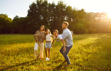 Fototapeta premium Happy young family of four having fun and playing plane at sunny day in nature. Parents mother, father and their children boy and girl walking in the field outdoors at sunset together.