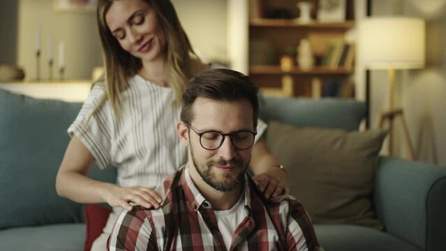 Smiling girlfriend massaging her boyfriend's neck while sitting on sofa at home