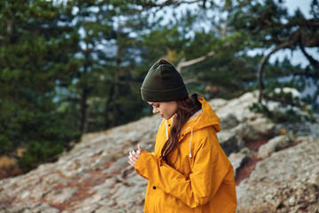 A woman in a yellow raincoat standing on the mountain peak checking her phone during a travel...