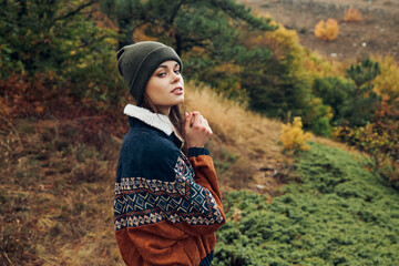 A young woman in stylish sweater and beanie standing on a picturesque hill during the autumn season