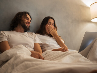 Peaceful sleepy couple in bed, resting against each other with a laptop nearby. Cozy nighttime...