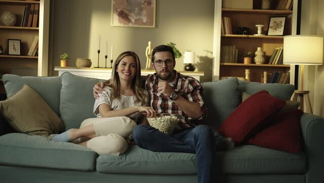 Young happy couple relaxing on sofa together, eating popcorn and watching movie. Shot in slow-motion