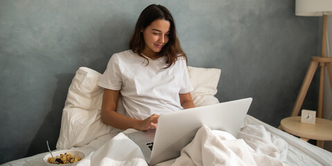 Young woman in bed during a video call on her laptop. Cozy and relaxed morning with breakfast...