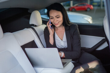 Happy young businesswoman typing at laptop and talking on smartphone while sitting on the back seat of car. Working online. Business, road trip, technology, travel concept