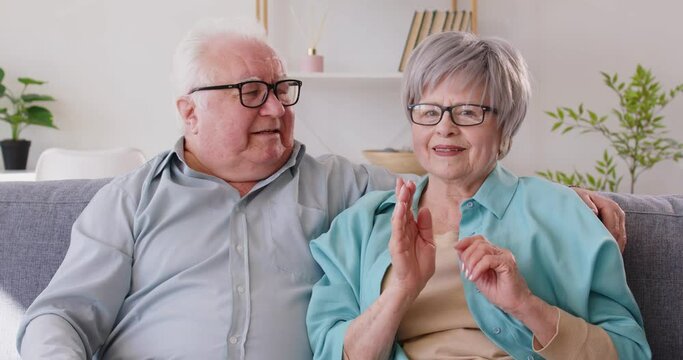 Happy old retired man and woman waving hand talking to webcam making online video call sitting on sofa at home. Senior couple enjoying webinar. Concept of internet communication. 4k video.