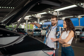 Smiling male instructor and young lady standing near car with open trunk on the parking. Driving...