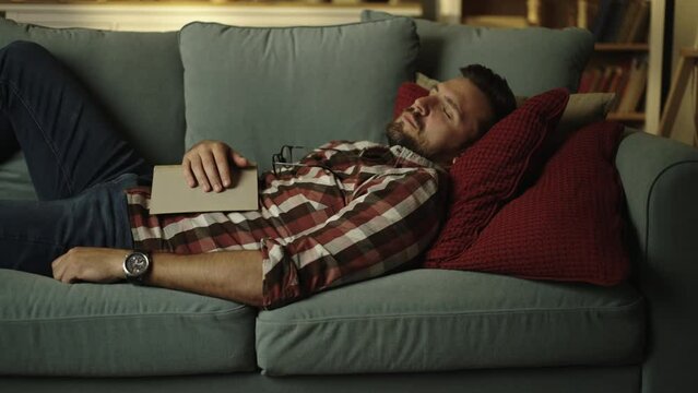 Young man with dark hair and beard is lying on couch and sleeping with a book on his chest