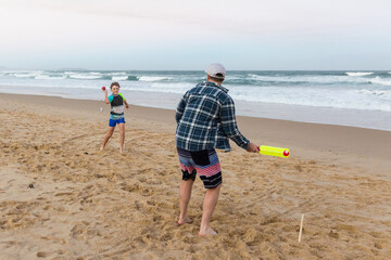 Boy throwing ball to his father holding bat playing cricket on the beach