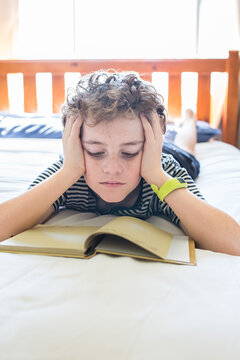 Boy lying on stomach on bed head resting in hands reading book natural light