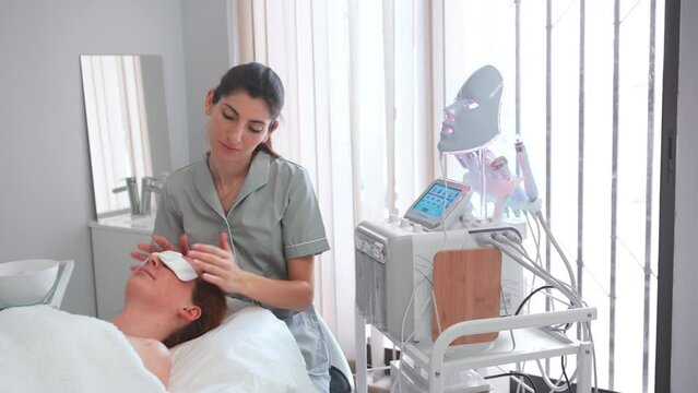 Female beautician professional using LED mask on woman patient for aesthetic treatment, relaxation, rejuvenation, color therapy, light therapy, skin care.