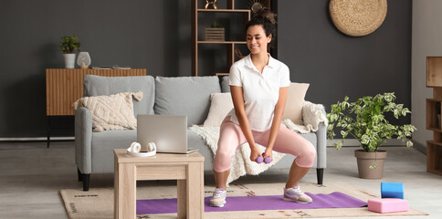 Sporty young African-American woman training with dumbbells in living room