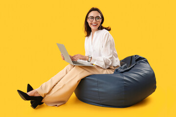 Pretty young woman working with laptop while sitting on bean bag chair against yellow background