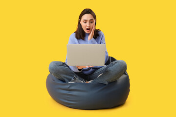 Surprised young woman with laptop sitting on bean bag chair against yellow background