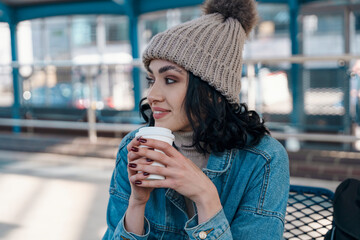 happy young woman drinking take away coffee and waiting for bus at bus station 