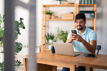 Happy smiling businessman wearing casual clothes and using modern smartphone in his home office at...