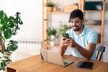 Happy smiling businessman wearing casual clothes and using modern smartphone in his home office, successful employer sitting at desk typing on mobile phone.