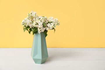 Vase with beautiful blossoming lilac on white wooden table against yellow background