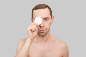 Man doing home skin care routine. Cotton pad near male eye. Dermatology treatment. Cream facial...