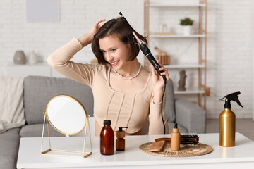 Beautiful young happy woman with curling iron and hair care products at home