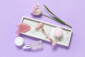Jade roller with gua sha, cosmetic cream, serum and tulip flower on lilac background