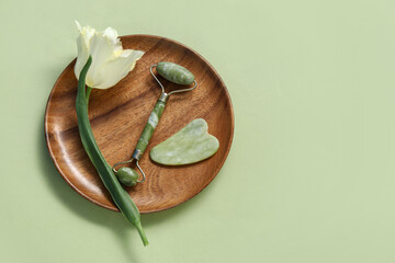 Wooden plate with jade roller, gua sha and tulip flower on green background