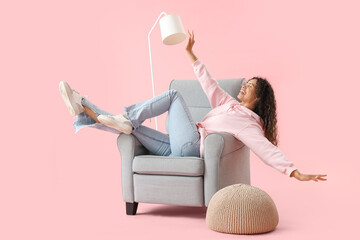 Young African-American woman lying in grey armchair on pink background