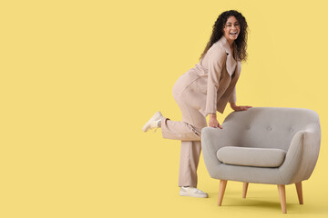 Young African-American businesswoman with grey armchair on yellow background