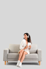 Fototapeta premium Young African-American woman in headphones with tablet computer sitting on grey sofa against light background