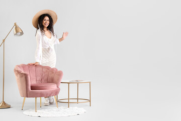 Young African-American woman with pink armchair, lamp and table on light background