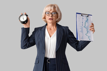 Stressed mature businesswoman with alarm clock and clipboard on light background