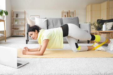 Sporty African-American woman with laptop and resistance band exercising on yoga mat at home