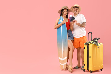 Happy young couple with suitcase, surfboard and passports on pink background