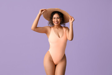 Beautiful young African-American woman in stylish swimsuit and wicker hat on lilac background
