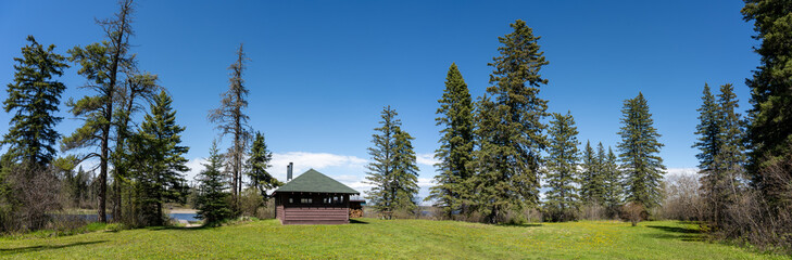 Panoramic view of a park picnic shelter surrounded by spruce and pine trees under a blue sky. A small lake can be seen through the trees.
